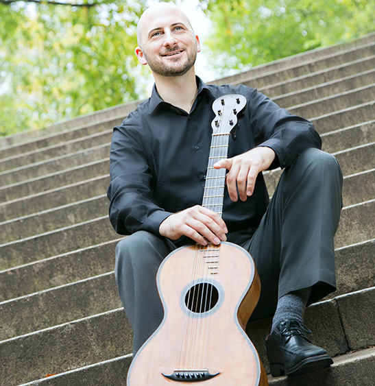 image of George England sitting on stairs outdoors, posing with a vertical guitar.