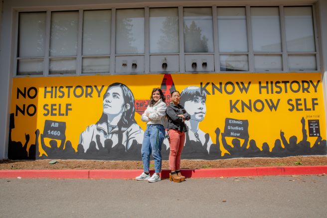 Two people posing in front of the AB 1460 mural on campus