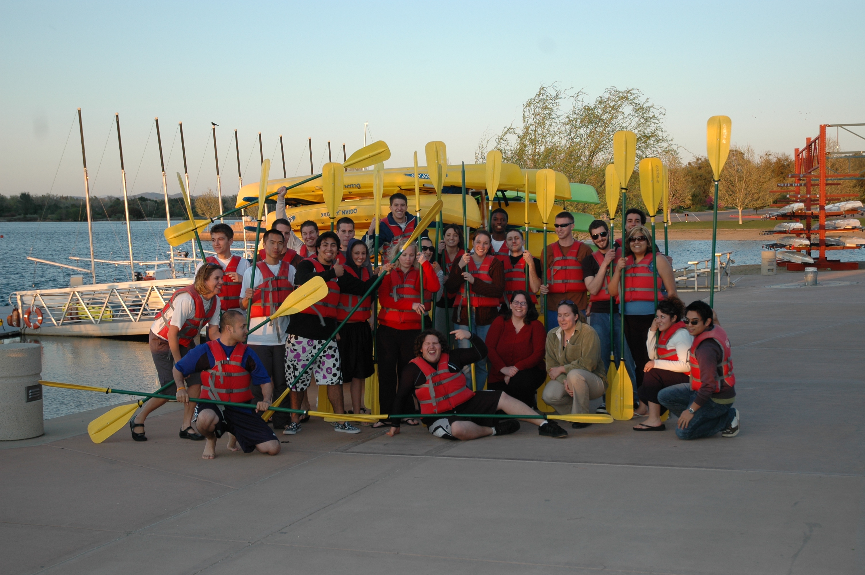 Students sitting in front of the CSUS Aquatic Center holding kayak oars and wearing life jackets