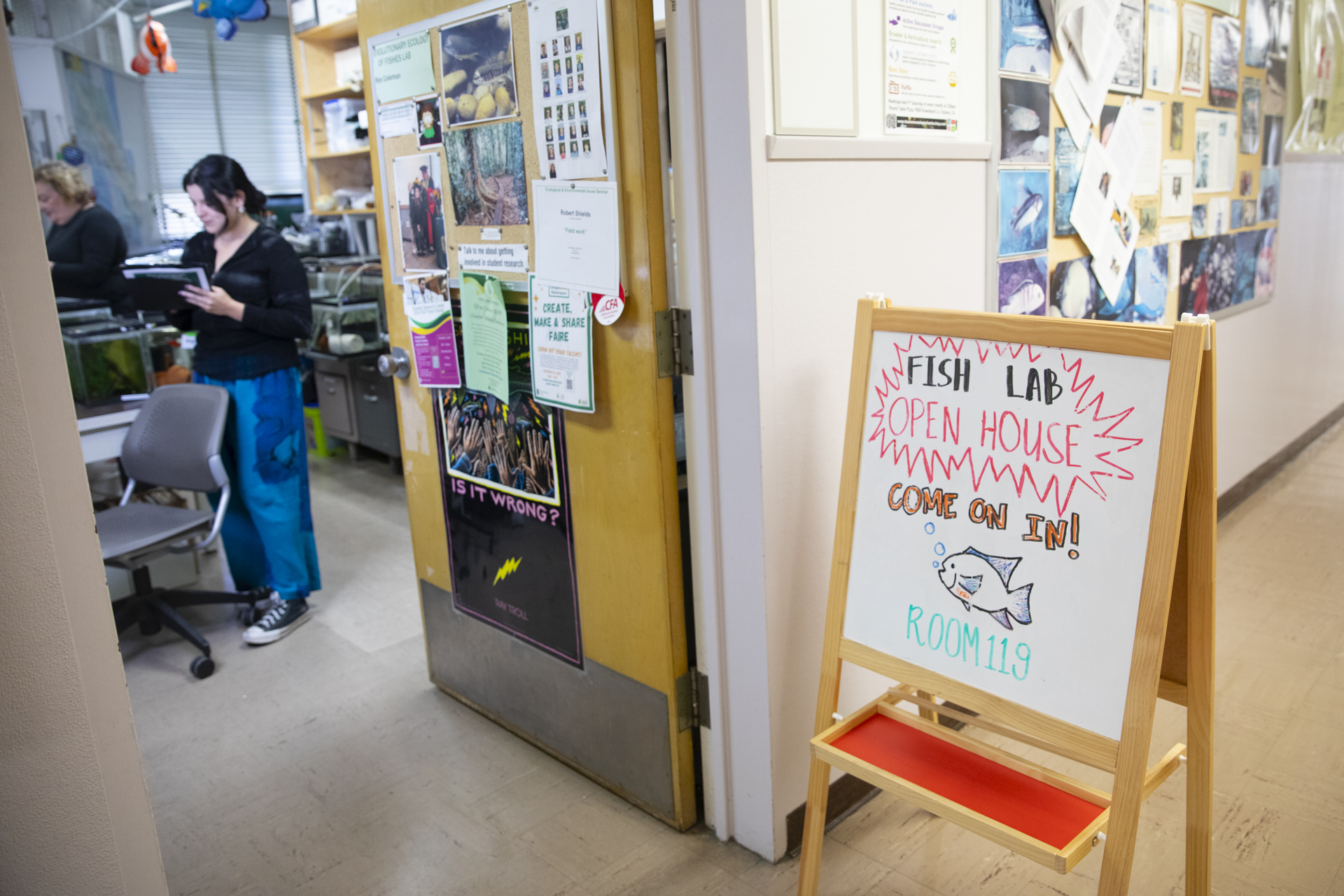 A sign reading "Fish Lab Open House, Come on in," pointing into a lab where a woman stands with a clipboard.